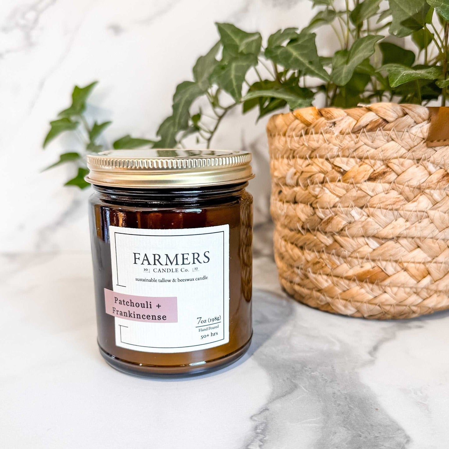Patchouli Frankincense scented candle by Farmers with natural soy wax in amber jar next to woven plant basket on marble surface