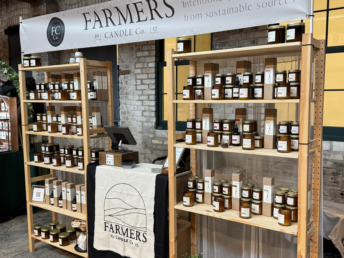 A market booth for Farmers Candle Co. displaying multiple wooden shelves filled with various scented candles in glass jars, with a white banner and branded towel hanging above.