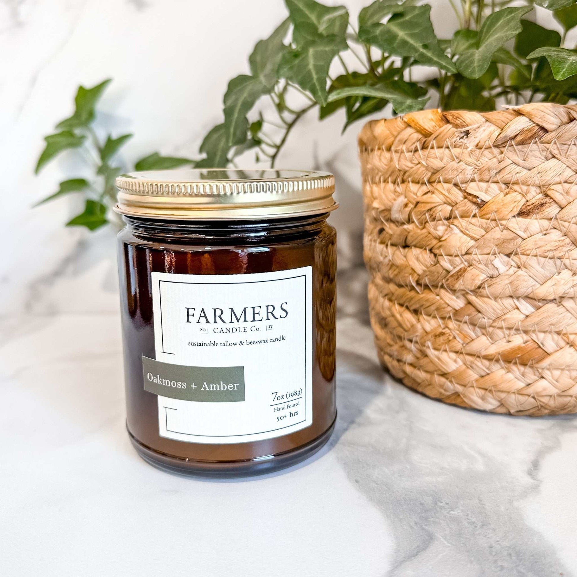 Oakmoss + Amber scented candle by Farmers Candle Co in amber glass jar with gold lid next to woven basket and green plant